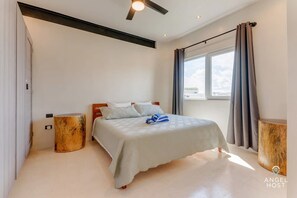 Une chambre à coucher avec un lit, un ventilateur de plafond, une fenêtre avec des rideaux et une petite table de chevet en bois.