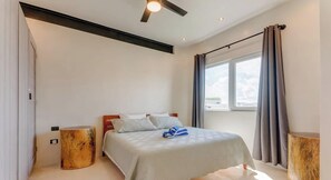 Een slaapkamer met een bed, een plafondventilator, een raam met gordijnen en een nachtkastje.
