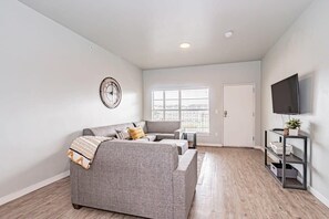 A modern living room with a grey sofa, a small table, a TV on a stand, and a window with blinds.