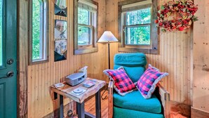 A cozy cabin interior with wooden walls, a teal armchair with checkered pillows, a small table with a lamp, and framed pictures on the wall. - Rustic Cabin w/ Patio & Pond on Blue Ridge Parkway (Ennice)