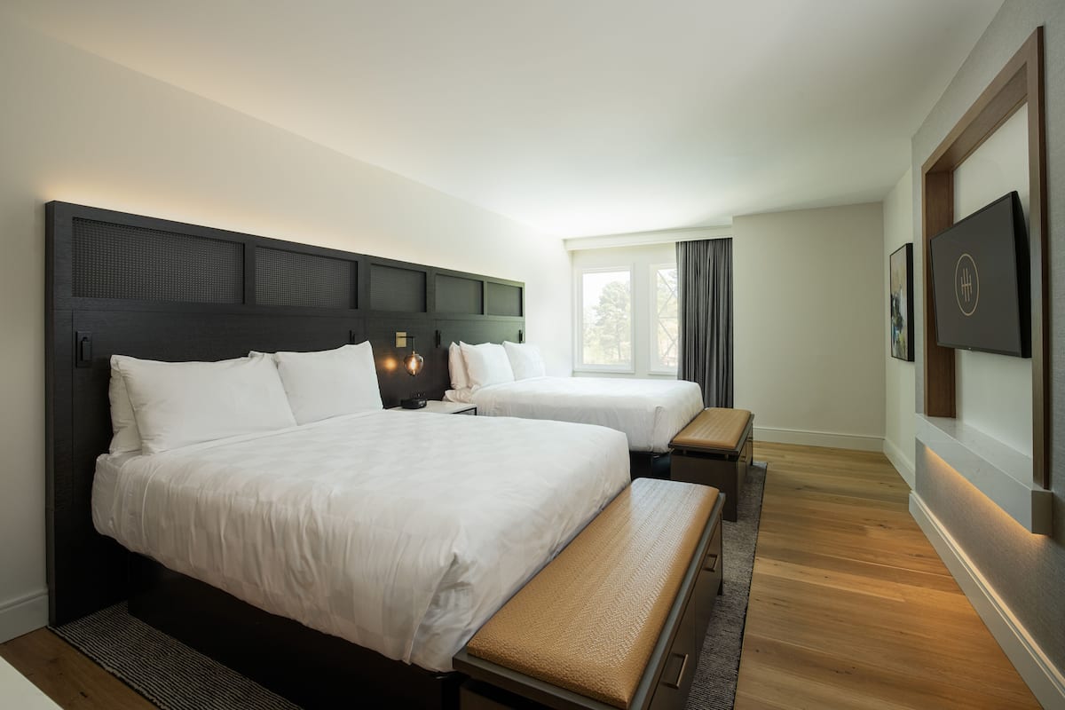 Ein Hotelzimmer mit zwei Betten, einer Sitzbank, einem großen Fenster und einem an der Wand befestigten Flachbildfernseher.