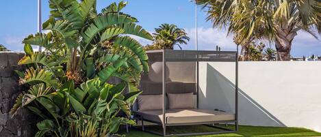 A poolside area with a sunbed, lush tropical plants, and a clear blue sky.