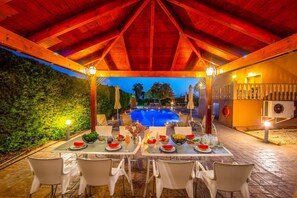 Un área de comedor al aire libre con mesas preparadas para una comida, con vistas a una piscina y una ladera boscosa.