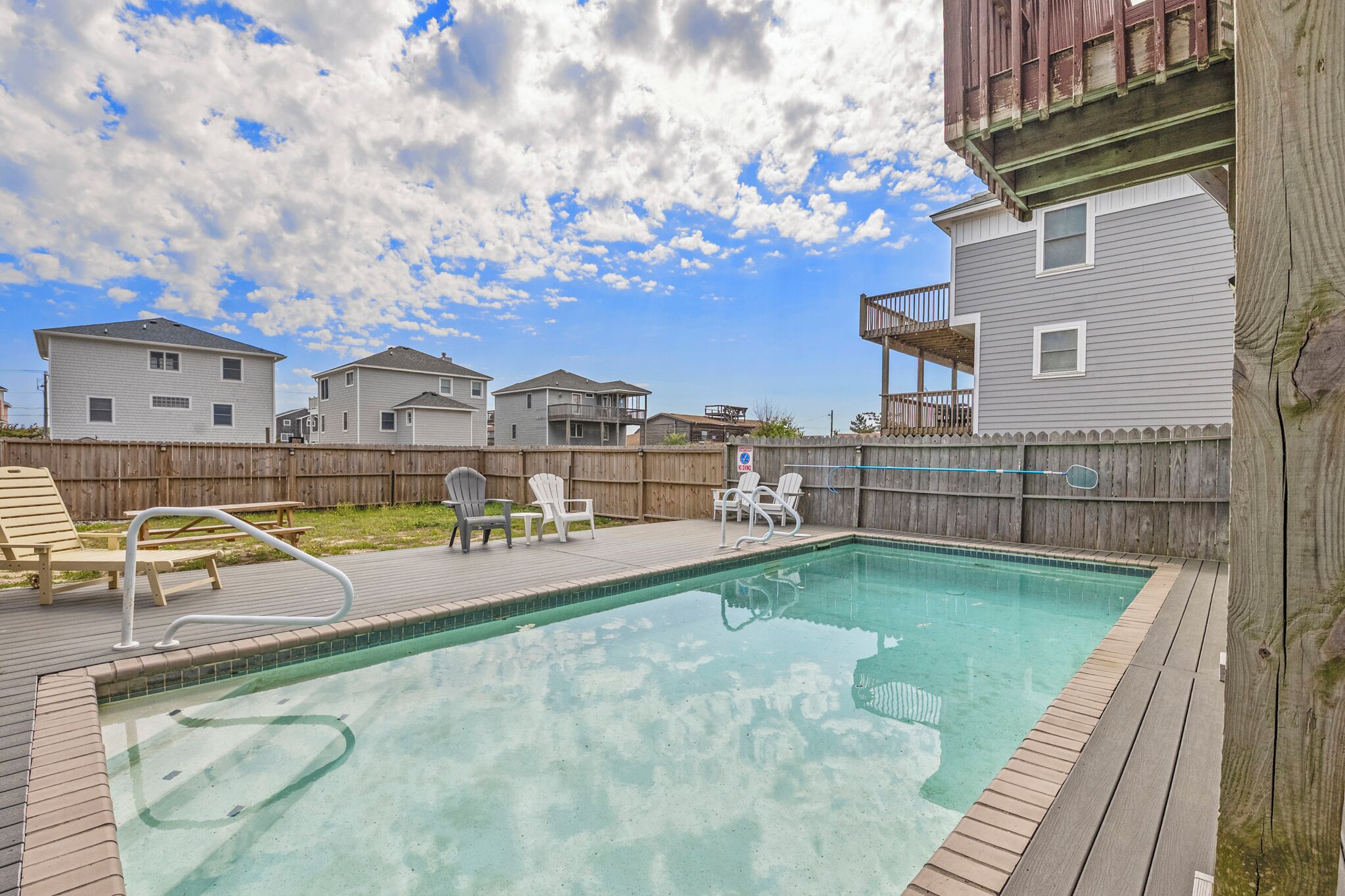 A backyard with a swimming pool, deck chairs, and a wooden fence.