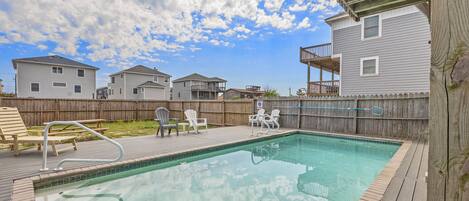 A backyard with a swimming pool, deck chairs, and a wooden fence.