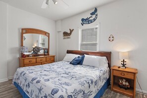 A bedroom with a bed, a dresser, a nightstand, a mirror, a window with blinds, and wall decorations. - Beach Bum'n: Private Pool | 3-Min Walk to Beach (Nags Head)
