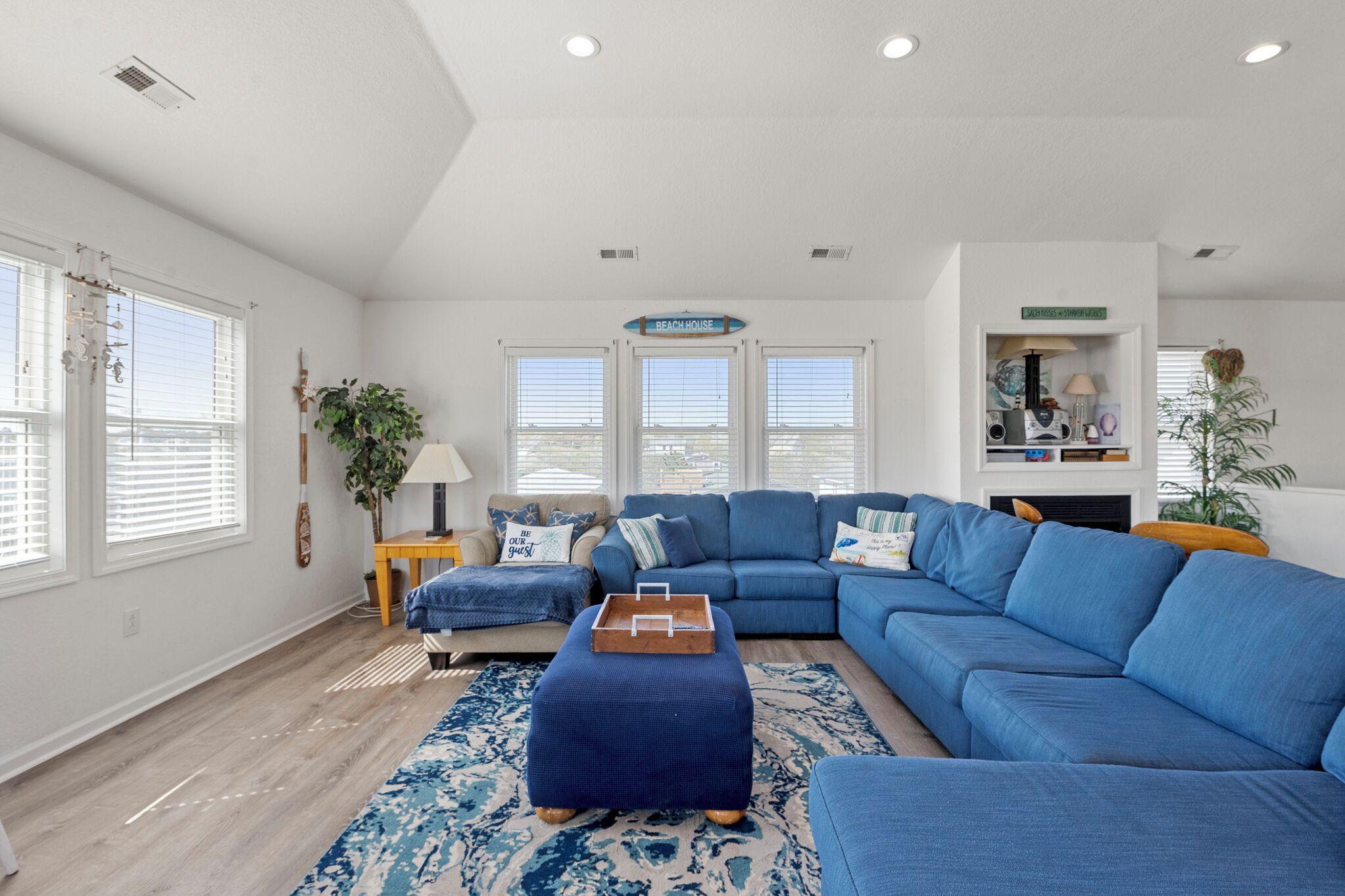 Una sala de estar con sofá modular, un reposapiés azul, alfombra estampada y un letrero que dice 'BEACH HOUSE'.