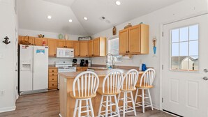 Una cocina con armarios de madera, un refrigerador blanco, un microondas y un lavamanos. Hay tres taburetes blancos en la isla.