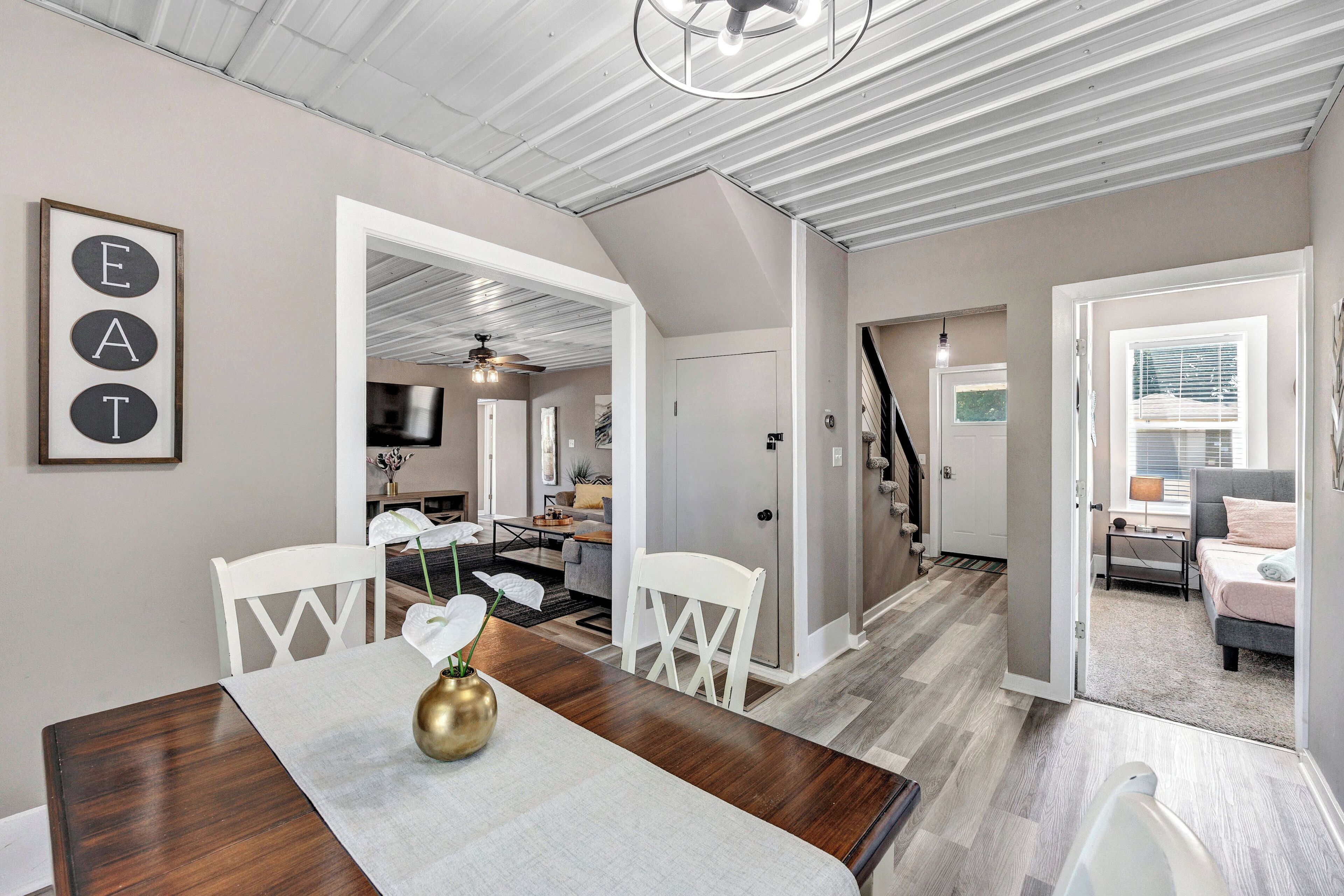 A modern interior with a dining area featuring a wooden table, white chairs, and a vase with flowers. There is a staircase leading to another level, and a hallway with a door and a small table.