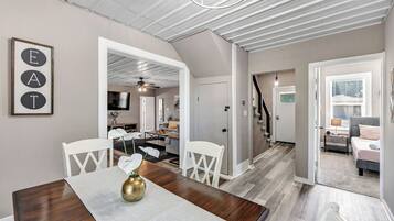 A modern interior with a dining area featuring a wooden table, white chairs, and a vase with flowers. There is a staircase leading to another level, and a hallway with a door and a small table.