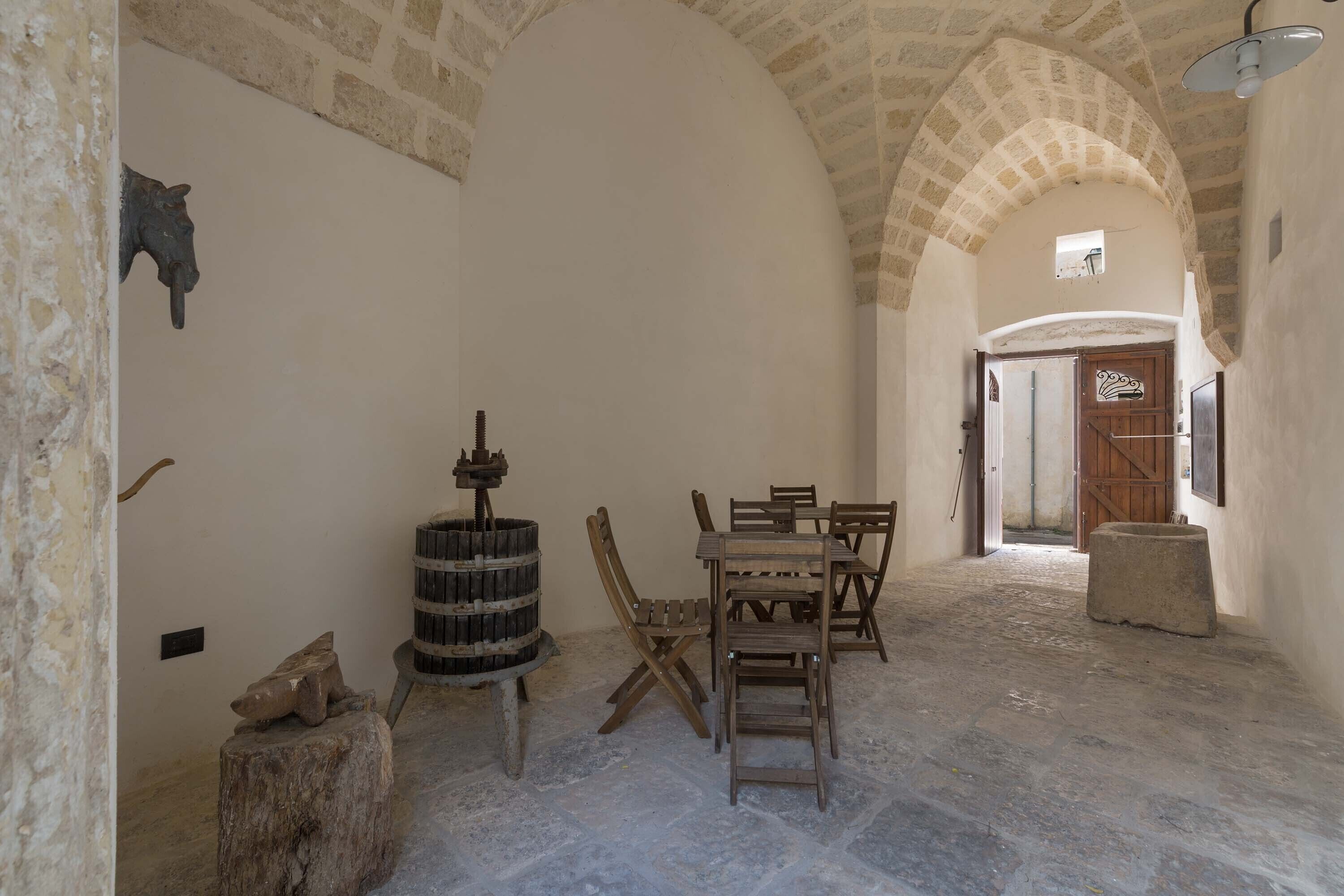 A narrow corridor with stone walls, a wooden table with chairs, and a barrel.