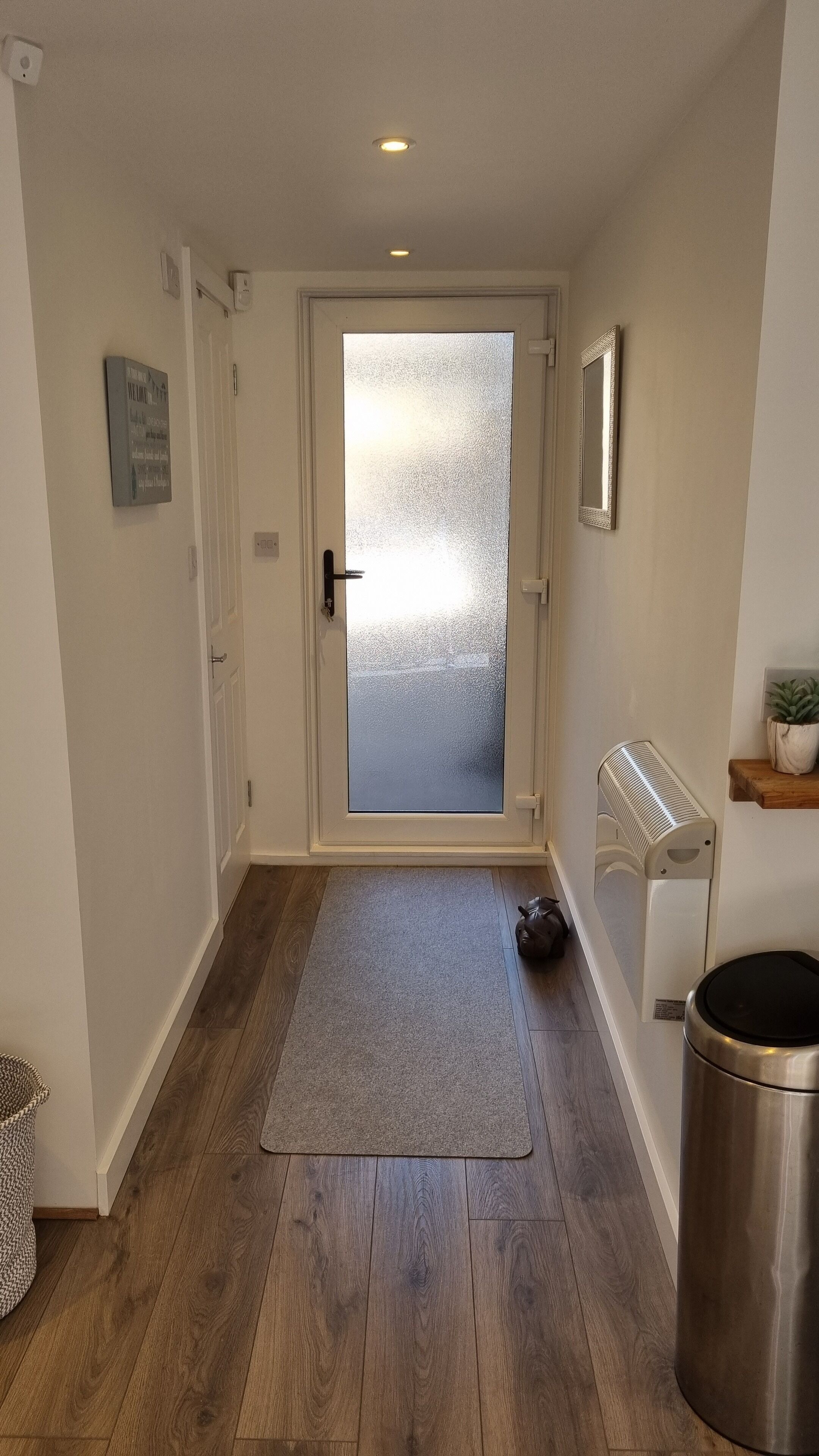 En hall med trägolv, en glasdörr, en matta, en papperskorg och en liten hylla med en växt.