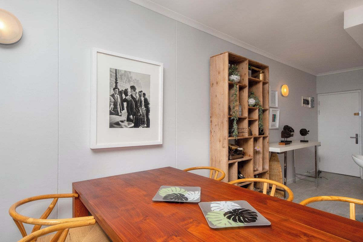 A dining area with a wooden table, chairs, a framed black and white photograph, and a wooden bookshelf with decorative items.