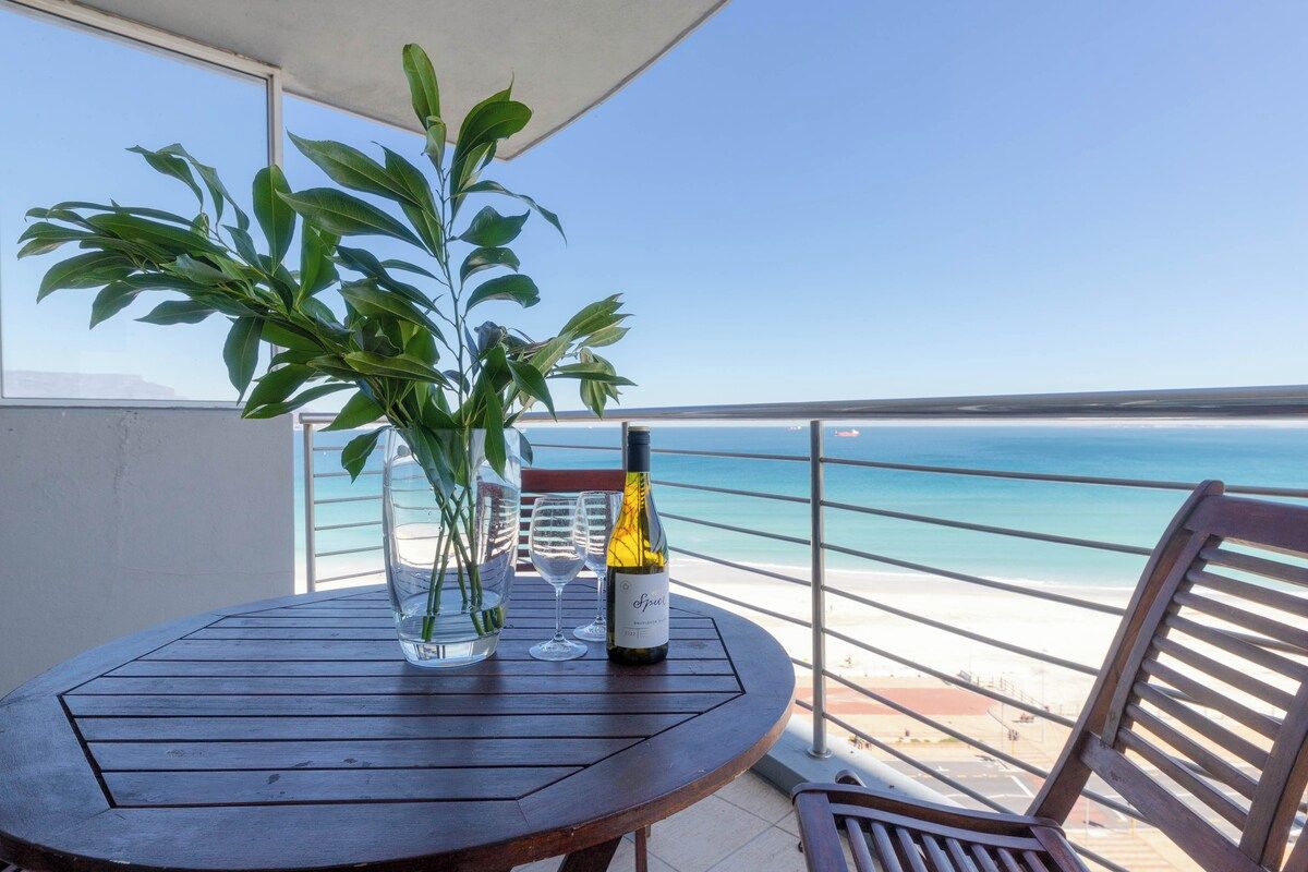 A round wooden table with a bottle of wine and glasses, a potted plant, and a view of the sea.
