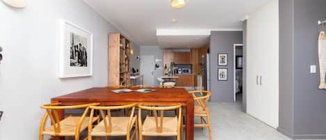 A dining area with a wooden table and chairs, a kitchen in the background, and a grey wall with a framed picture.