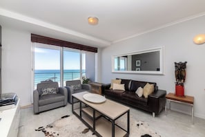 A modern living room with a sofa, armchair, and coffee table, a large window with a view of the sea, and a mounted artwork on the wall. - West Coast Horizon Retreat (Cape Town)