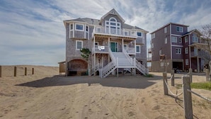 Une propriété en bord de mer, composée de plusieurs étages, avec un escalier imposant menant à l’entrée principale, un espace extérieur couvert et une plage de sable en premier plan.