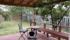 Una terraza techada al aire libre con una mesa y dos sillas, rodeada por un cerco de madera y árboles.