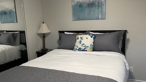 Una cama bien hecha con colcha blanca y gris, un cojín floral y una mesita de noche con una lámpara. Hay un cuadro en la pared sobre la cama.