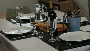 A dining table set with plates, glasses, and a pitcher. There is a television in the background and a window with blinds.