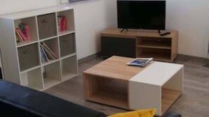 A modern living room with a black sofa, a wooden coffee table, and a TV on a wooden stand. There is a bookshelf with books and a painting on the wall.