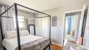 A four-poster bed with a grey bedspread, a bedside table with a lamp, and a view of a hallway with a door leading to another room. - Spacious 3 Bedroom, 5 minutes from Downtown Cincinnati (Highland Heights)