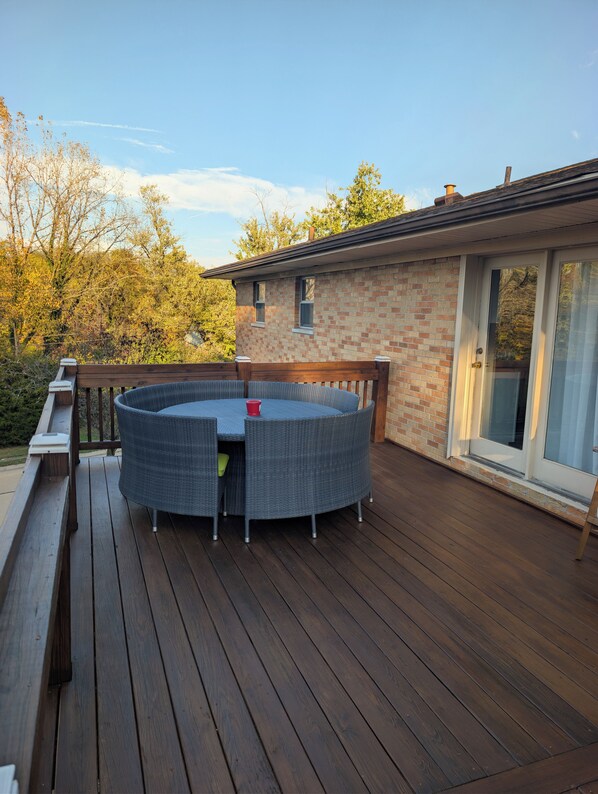 A wooden deck with a round wicker table and chairs, a glass door, and a brick building in the background.