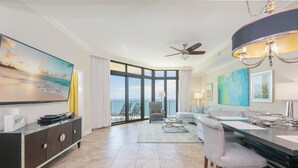 A modern hotel room with a large window, a ceiling fan, a dining area, and a view of the ocean. - The Oasis at Orange Beach, formerly known as Phoenix West II, Unit 2103 (Orange Beach)