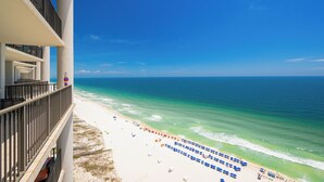 A beach view from a balcony with a clear sky, ocean, and beach umbrellas. - The Oasis at Orange Beach, formerly known as Phoenix West II, Unit 2103 (Orange Beach)