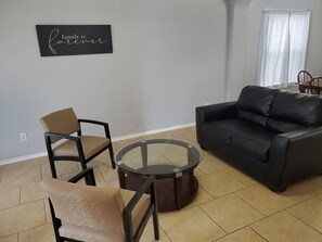 Un salon avec un canapé en cuir noir, deux fauteuils beiges et une table basse en verre. Un panneau accroché au mur porte l’inscription : « La famille, c’est pour toujours. »