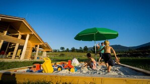 A sandy play area with a green umbrella, wooden play structures, and a building in the background. - Quadruple Room with Bathroom (Sciegny)