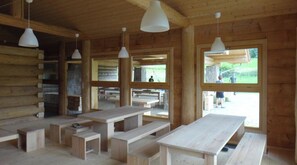 A wooden cabin interior with multiple wooden benches and tables, large windows, and hanging pendant lights. - Quadruple Room with Bathroom (Sciegny)