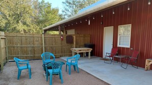 A covered patio area with a wooden table and blue plastic chairs, a red folding chair, and a red building with a window and string lights. - Country Living with Kyle Field 30 minutes away (SOMERVILLE)