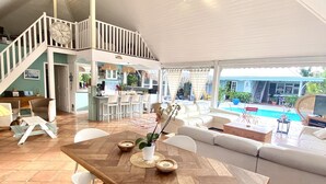 Un vaste espace de vie comprenant une table à manger en bois, des chaises blanches et un canapé blanc. Un escalier mène à l’étage supérieur.