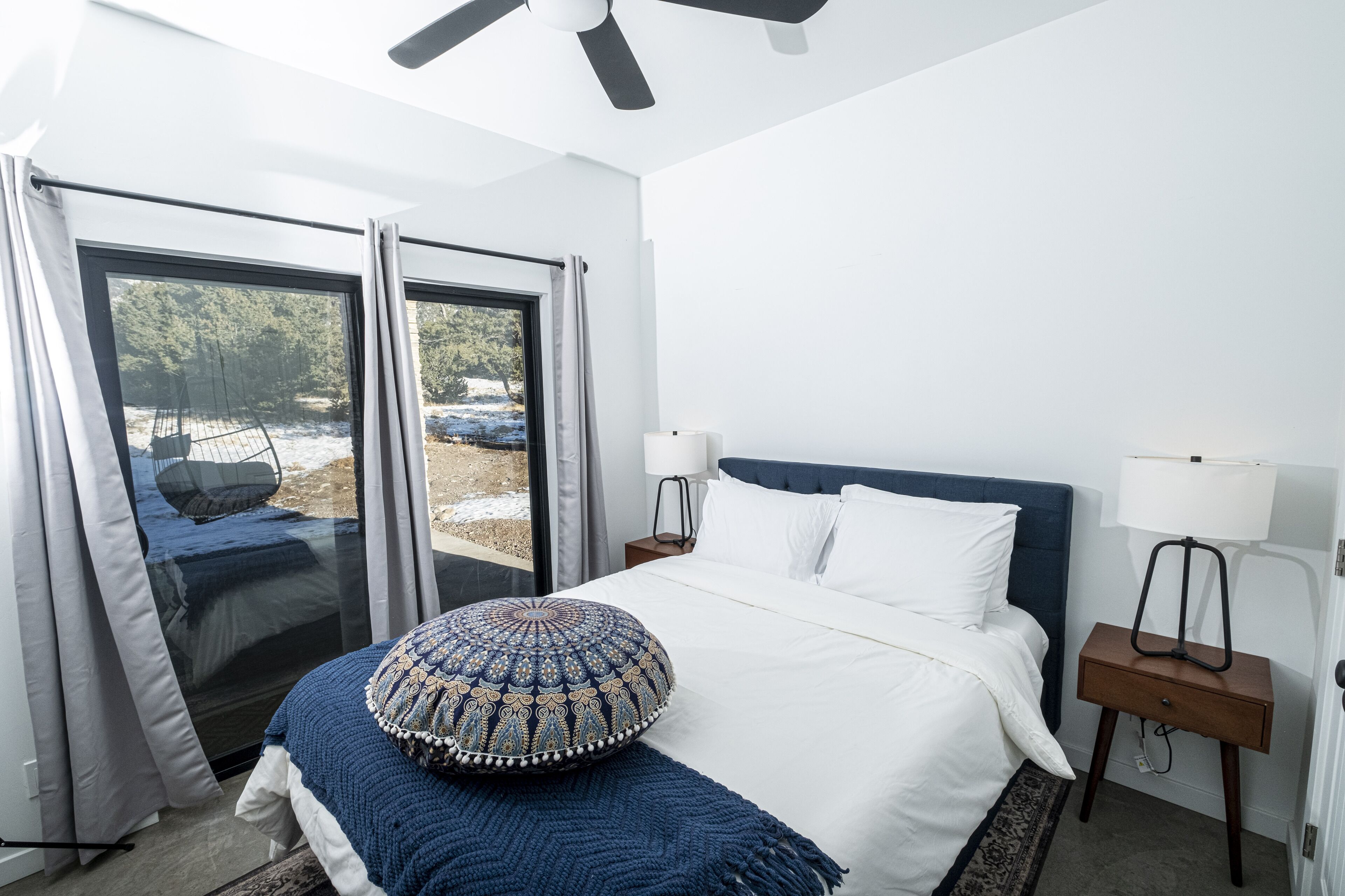 A bedroom with a bed, two bedside lamps, a nightstand, a window with curtains, and a ceiling fan.