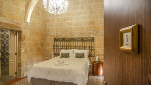 Un dormitorio con pared de piedra, una araña, una cama con ropa de cama blanca y una puerta de madera con una placa con número.