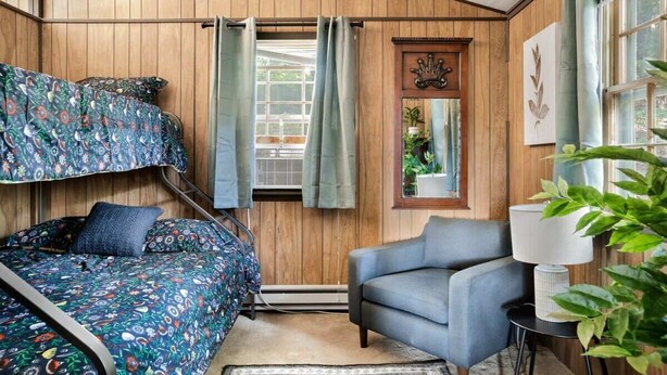 A room with a bunk bed, a sofa, a small table with a lamp, and a window with curtains. - Secluded Cabin: Hot Tub, Huge Deck, Fire Pit, WiFi (Hedgesville)