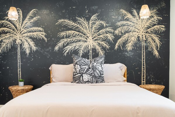 A bed with white bedding, two black and white patterned pillows, and a black wall with a palm tree mural.