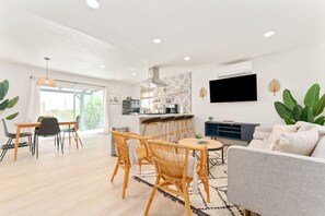 A modern living room with a sofa, dining table, and chairs, a kitchen area with bar stools, and a large TV mounted on the wall.