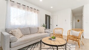 Ein Wohnzimmer mit einer Couch, zwei Korbstühlen, einem runden Tisch mit einer Pflanze und einem gemusterten Teppich.