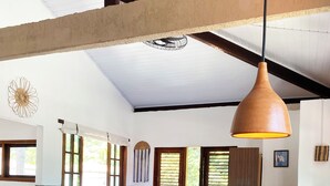 A room with a wooden table, a sofa, a bed with a blue bedspread, and a hanging pendant light. - Santo Hostel (Porto Seguro)