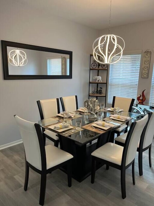 A dining area with a glass table surrounded by white chairs, a black chandelier, and a mirrored wall.