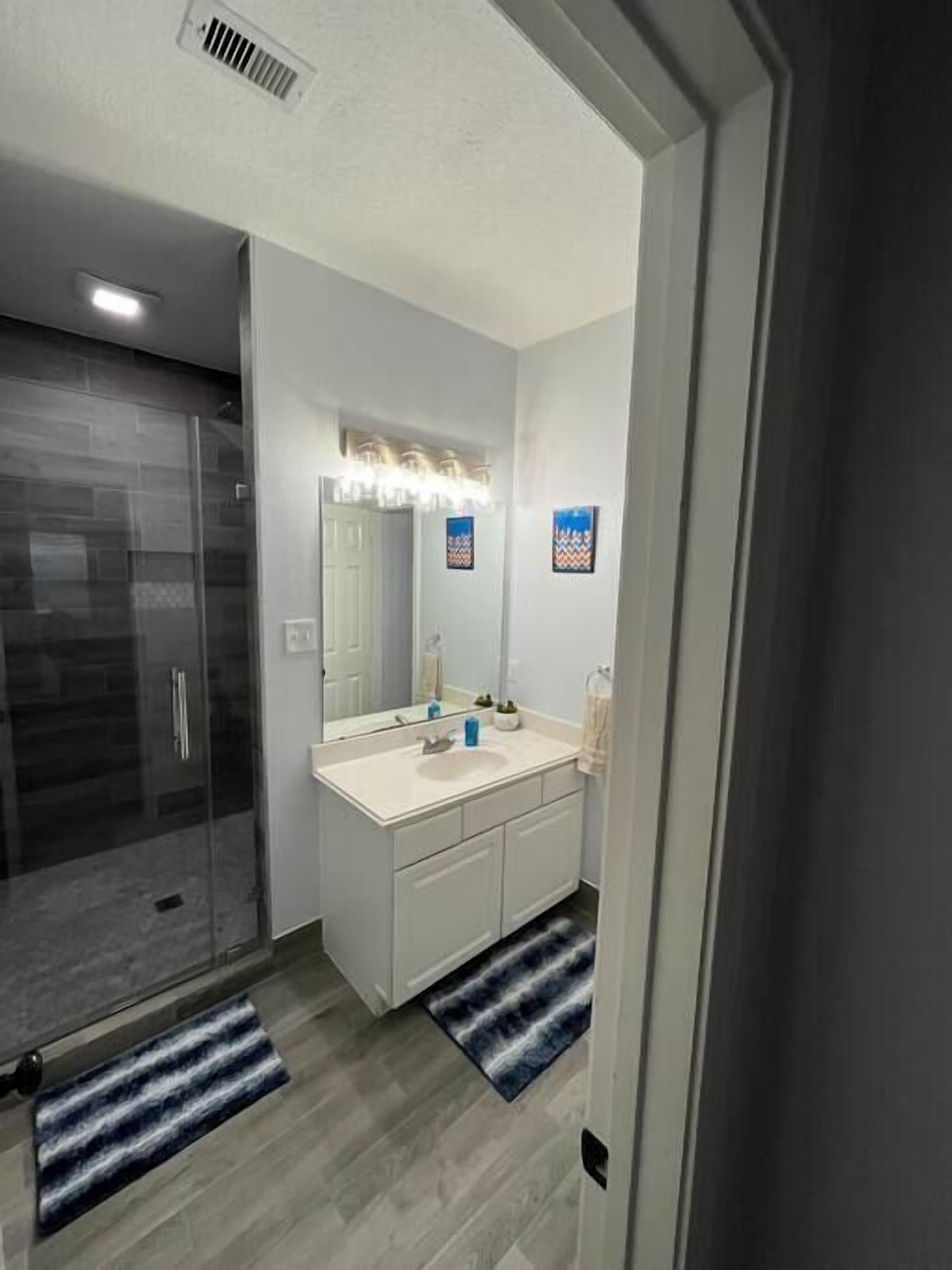 A modern bathroom with a glass shower enclosure, a white vanity with a mirror, and two blue and white striped rugs.
