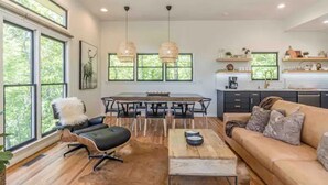 Uma sala de estar moderna com sofá, mesa de centro e área de jantar com mesa e cadeiras. Grandes janelas oferecem luz natural e vista para o verde lá fora.