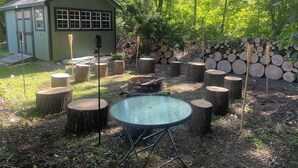 Un área de estar al aire libre de estilo rústico, con una mesa redonda de vidrio rodeada de taburetes de madera y un cobertizo pequeño.