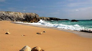 Uma praia arenosa com pedras grandes e um paredão rochoso ao fundo.