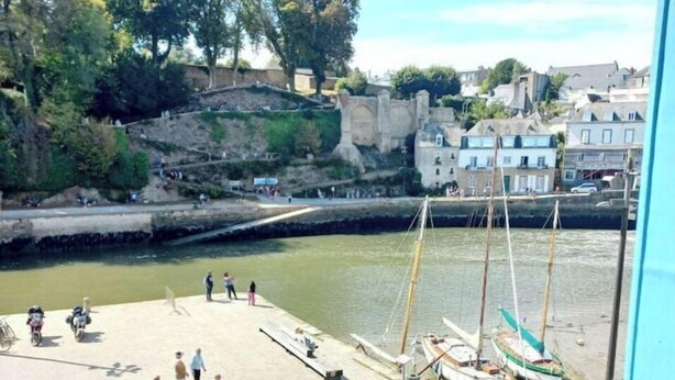 Um calçadão à beira do rio com mesas e cadeiras, barcos e um muro de pedra ao fundo.