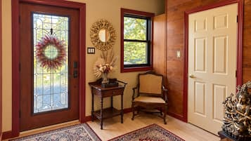 Un vestíbulo con puerta de madera, puerta de cristal adornada con una corona, una mesita con un florero, una silla de mimbre y un gran panel de pared de madera.