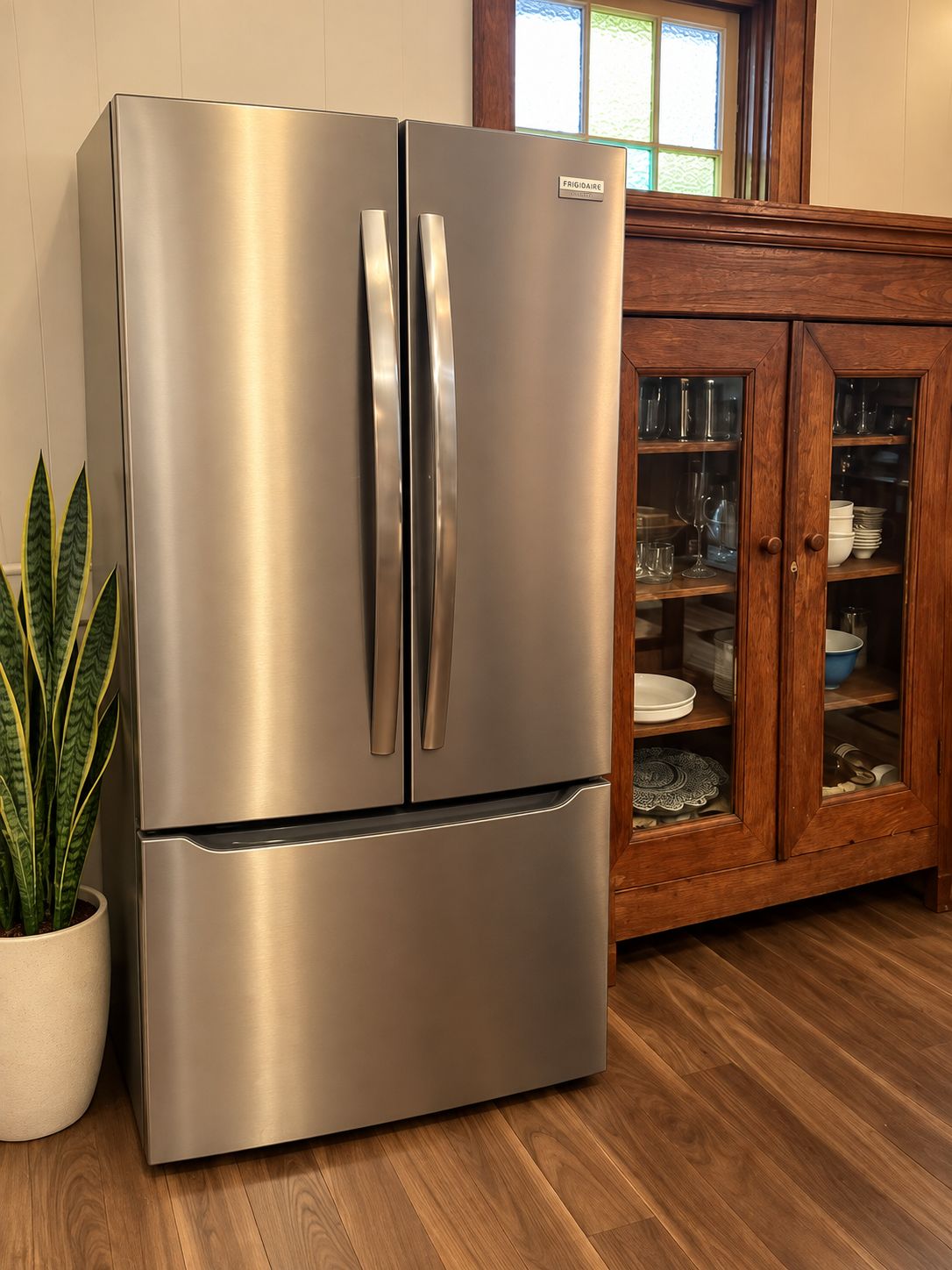 Ein edelstahlfarbener Kühlschrank mit zwei Türen, ein Holzschrank mit Glastüren und ein Blumentopf.