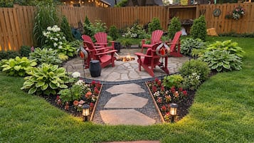 Une terrasse à l’arrière de la maison, avec des chaises Adirondack rouges, un chemin de pierres et une variété de plantes.